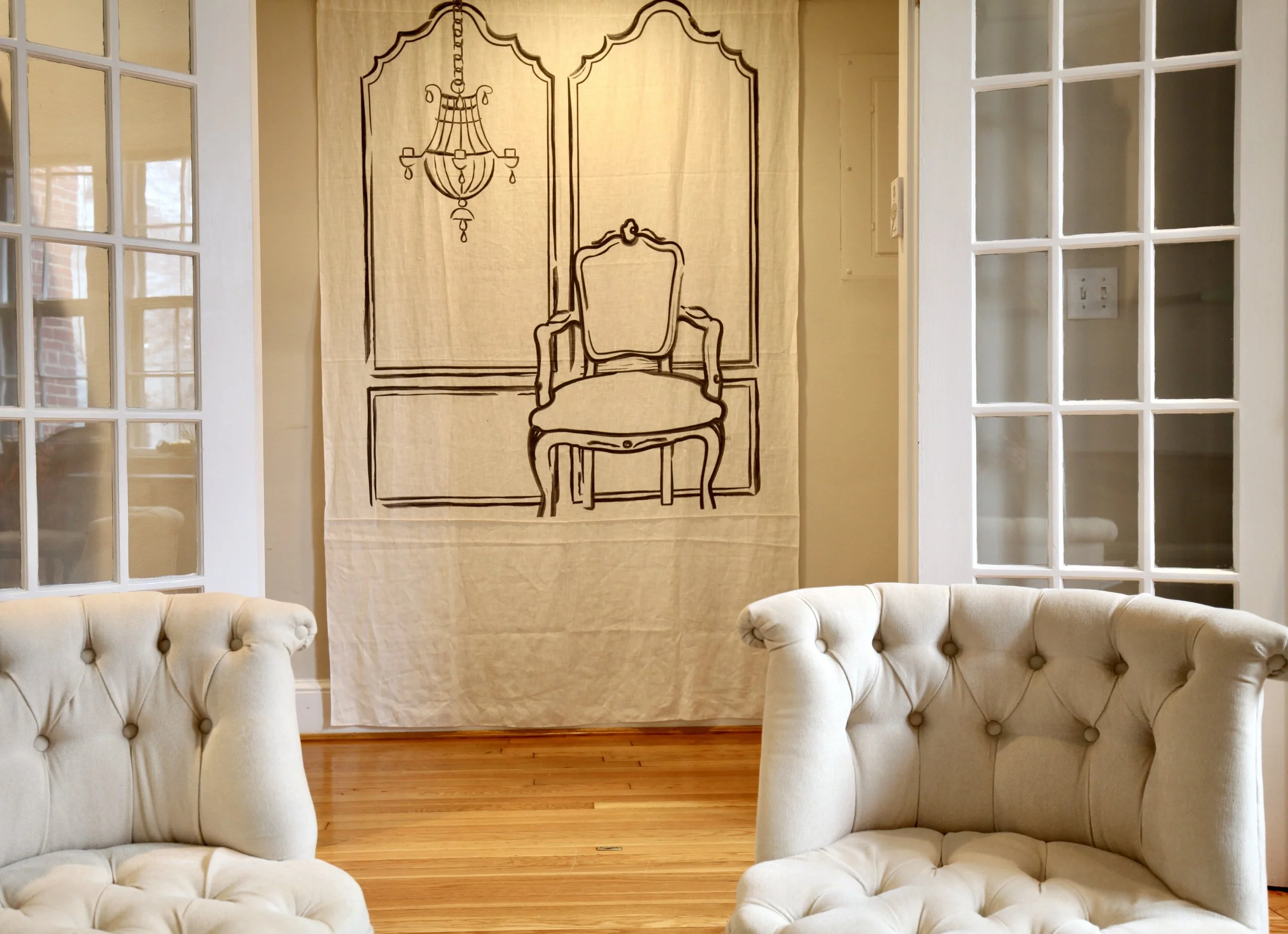 Interior of a living room with beige tufted armchairs, a hanging fabric wall art with a sketch of a chair, chandelier, and wall panels, french doors and hardwood floor.