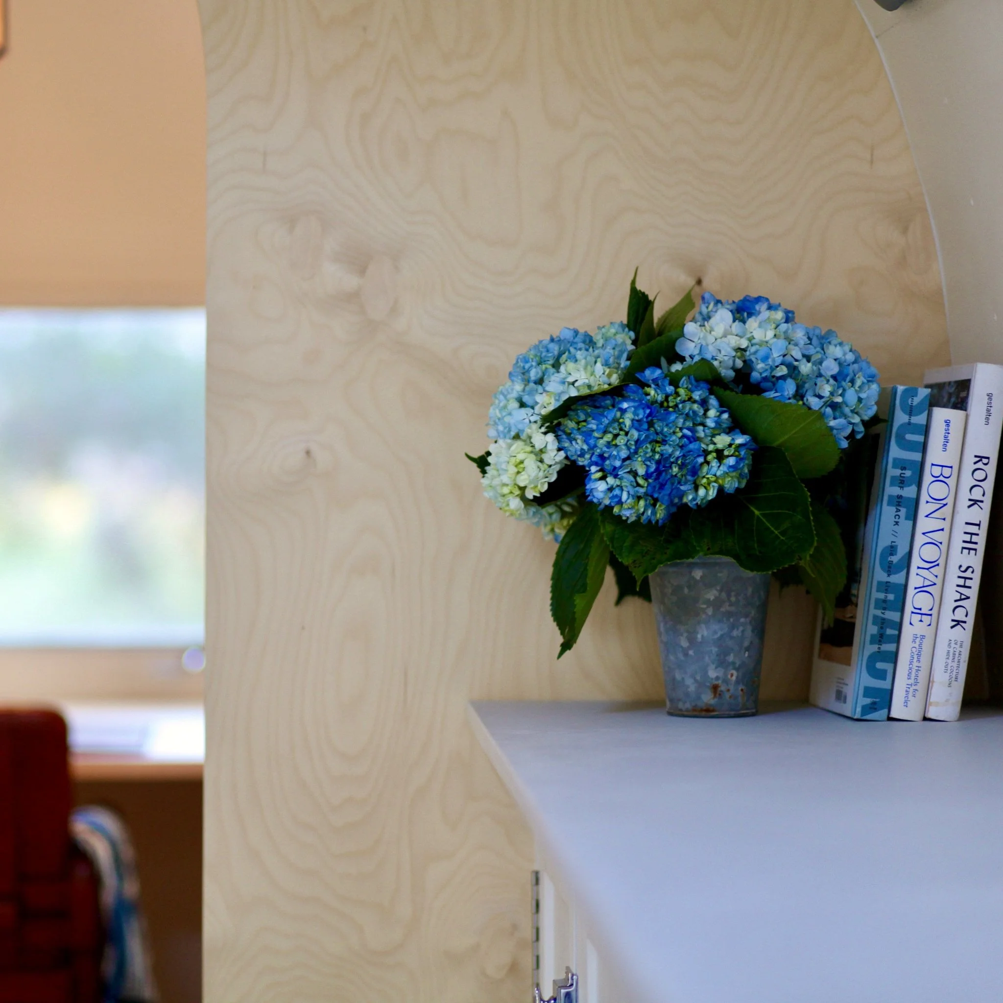 A bouquet of blue and white hydrangeas in a gray textured vase placed on a white shelf next to a stack of books on a light wooden side table.