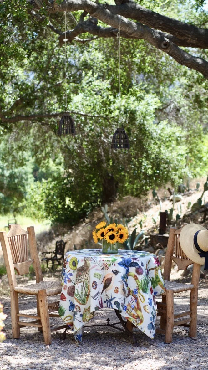 A table with a colorful, nature-themed tablecloth featuring animals and plants, set outdoors under a tree, with a bouquet of sunflowers, two wooden chairs, and a wide-brimmed hat hanging on one of the chairs.