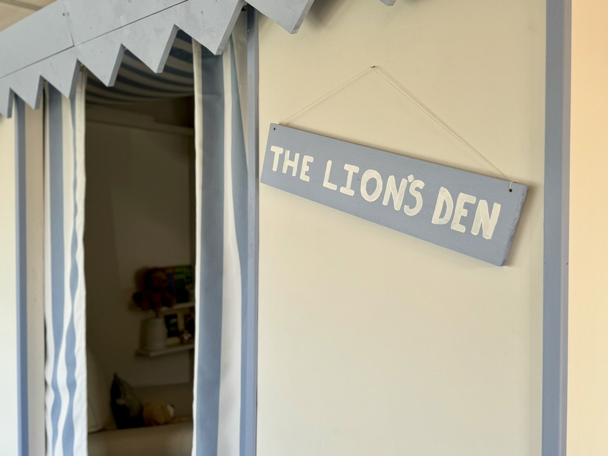 Interior corner of a play area with a sign reading 'The Lion's Den' hanging on the wall and a small section of soft play area with a curtain and some stuffed animals.