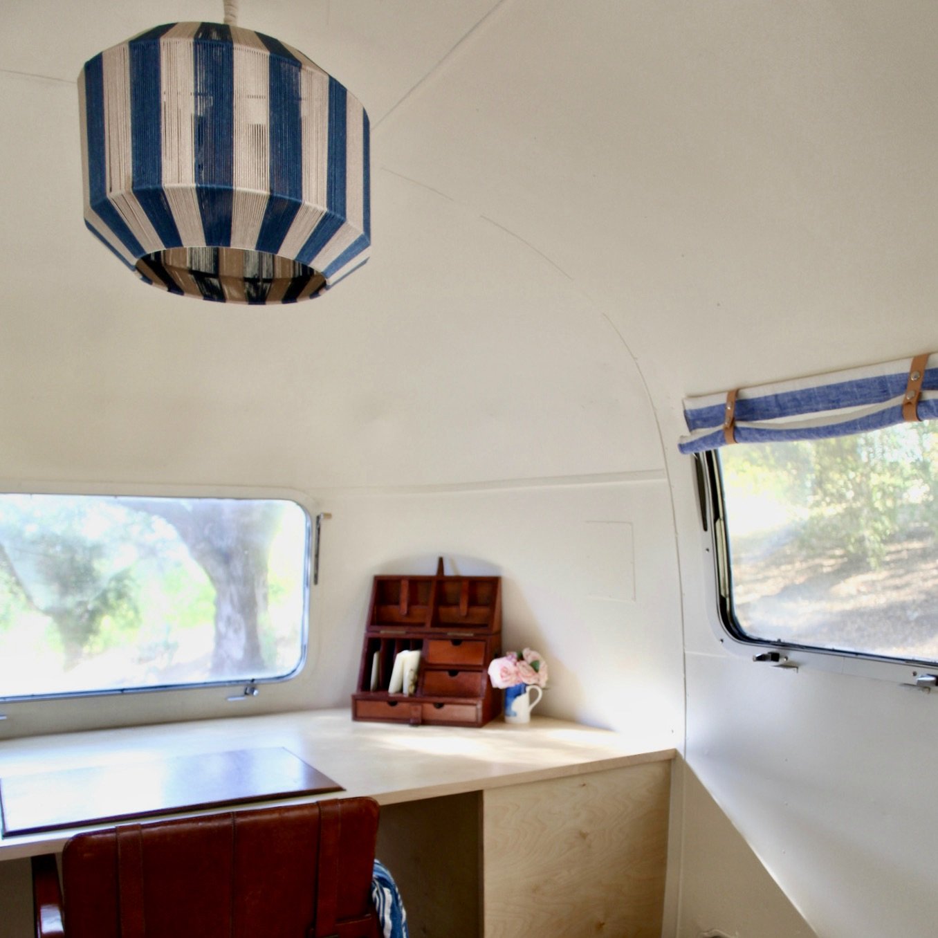 Interior of a cozy camper with white walls, a small window with blue striped curtains, a wooden workspace with a small wooden organizer, a pink flower bouquet, and a brown leather chair. A blue and white striped paper lantern hangs from the ceiling.