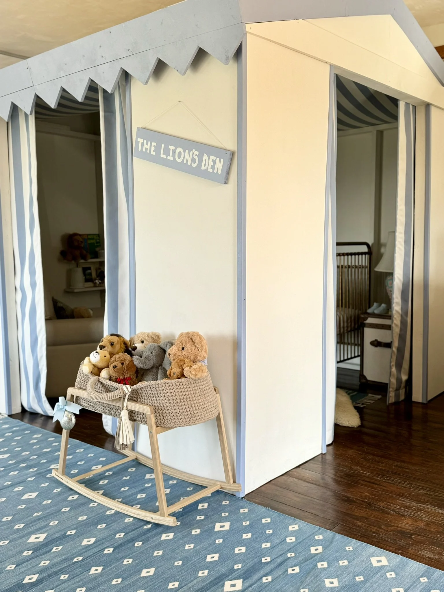A toy tent labeled "The Lion's Den" with a group of plush stuffed animals and a wooden rocking cradle in front of it, situated on a blue patterned rug in a playroom.