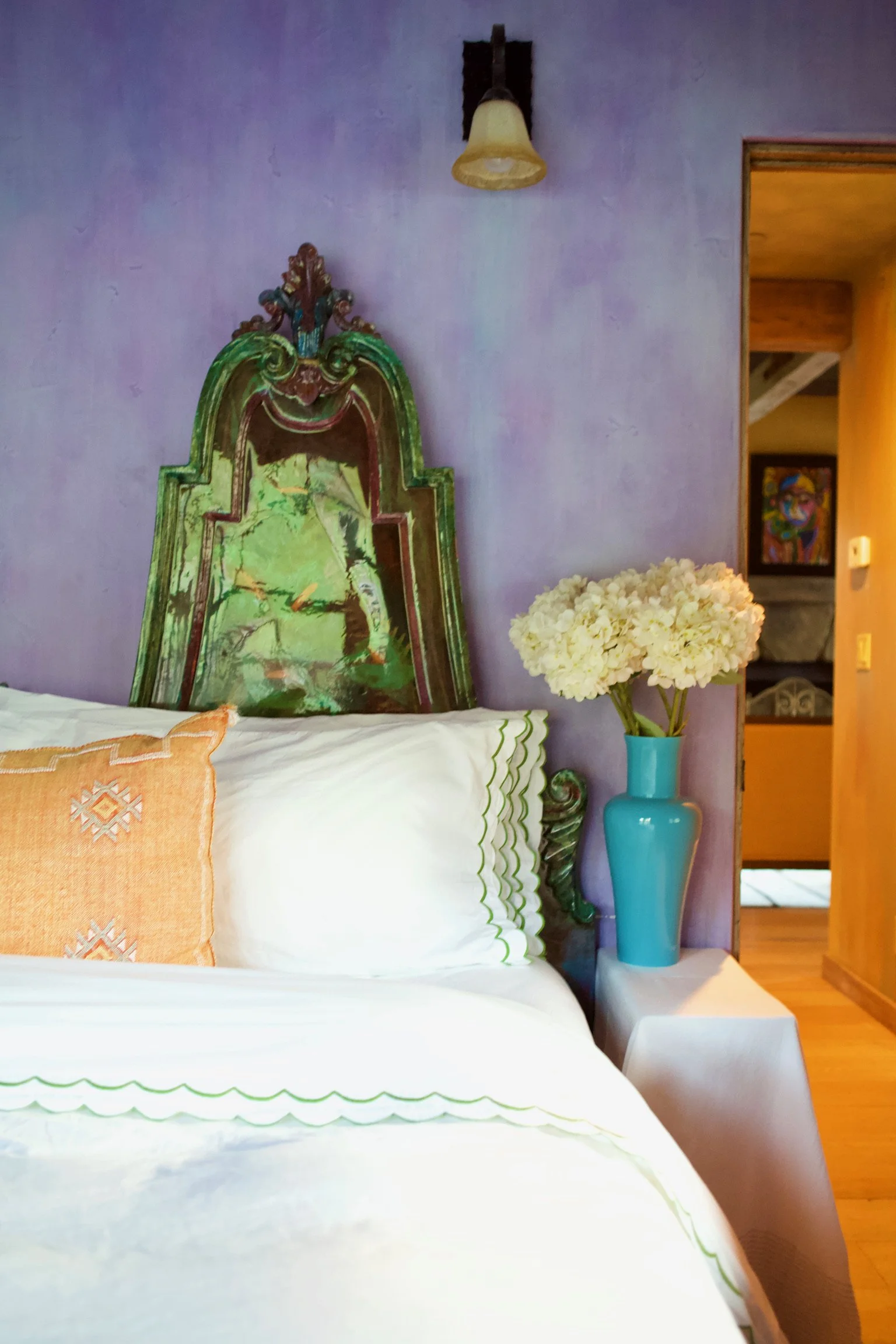 A bed with white linens and an orange embroidered pillow, a side table with a turquoise vase filled with white hydrangeas, a purple wall with an ornate green and brown wooden headboard, and a doorway showing a colorful framed art piece inside a cozy 