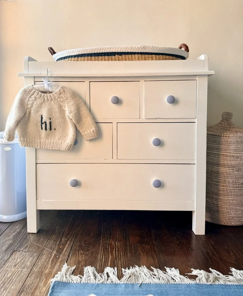 White dresser with five drawers, with a small cream-colored sweater hanging on a hook on the left side, and a woven basket on the right side. On top of the dresser, there is a fabric-covered changing pad. The floor is dark wood, and a fringed rug wit