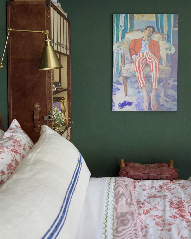 A cozy bedroom with a green wall, a wooden headboard with a brass reading lamp, white bedding with red floral patterns, and a colorful portrait of a woman in striped pants hanging on the wall.