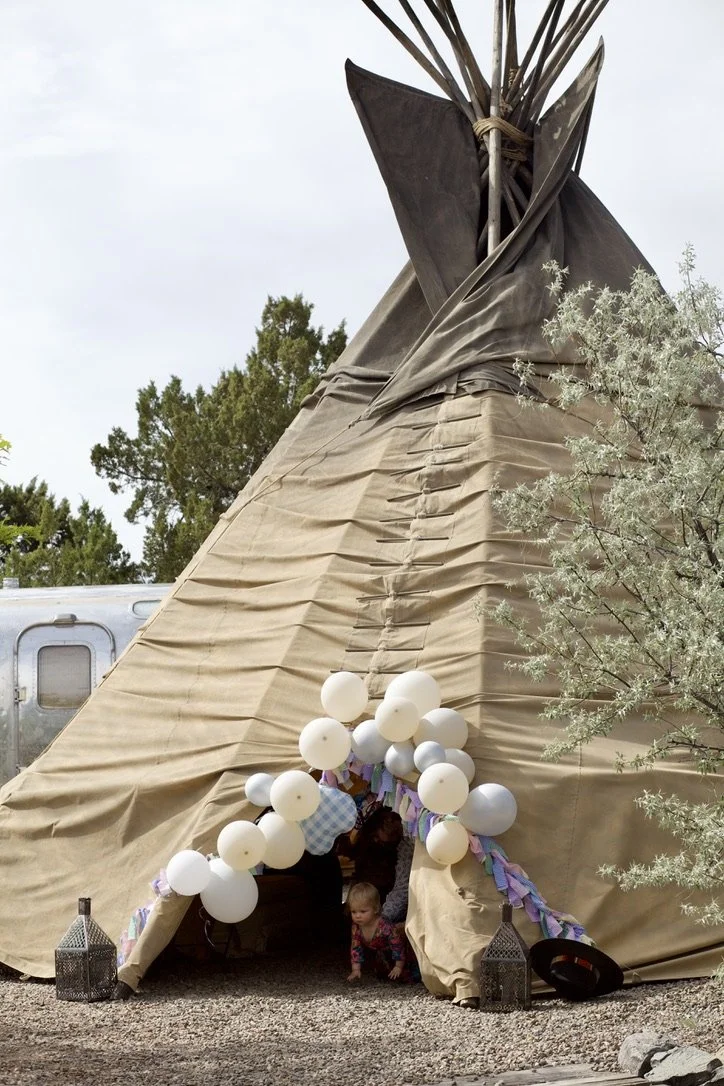 Front view of a large beige teepee decorated with white balloons and colorful fabric strips, with children sitting at the entrance, outdoors with trees and a silver trailer in the background.