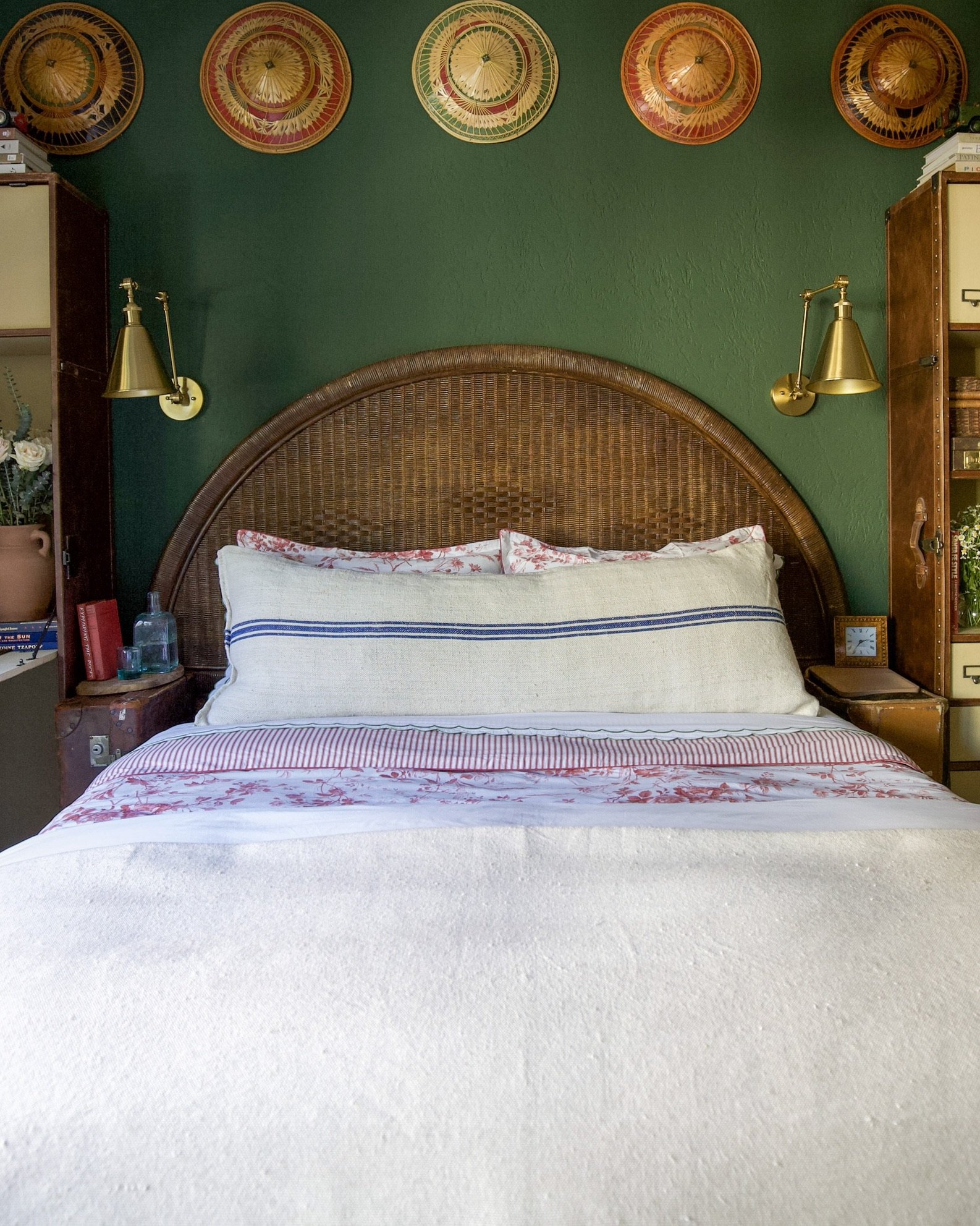 A bed with a wicker headboard against a green wall, decorated with five colorful paper umbrellas, flanked by gold wall-mounted lamps, with a nightstand on each side.