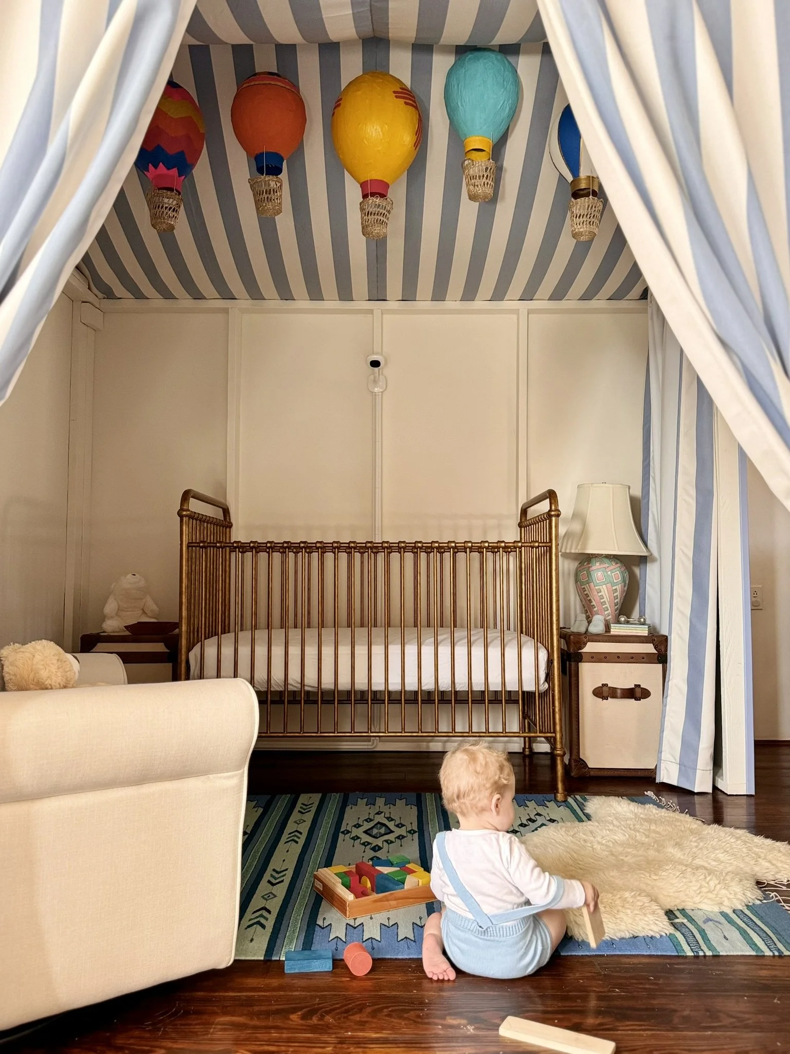 A nursery with a striped blue and white canopy and hanging colorful hot air balloon decorations on the ceiling. There is a small child sitting on a rug in front of a shepherd's cot, playing with toys. The room has a beige armchair, a nightstand with 