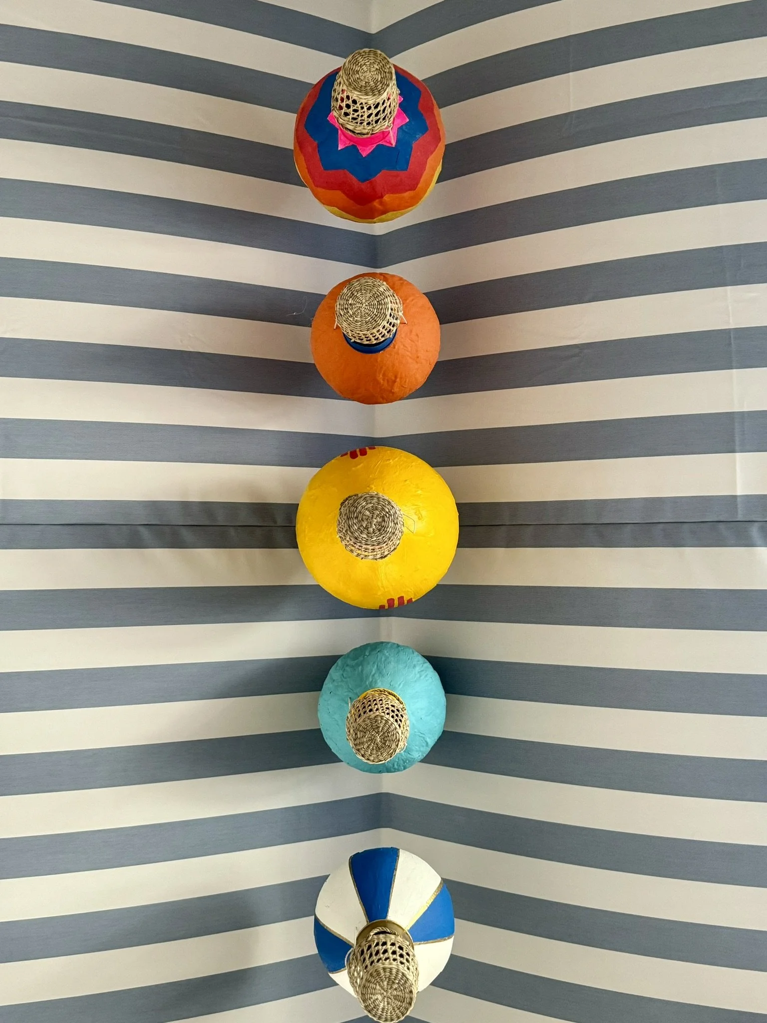 Five colorful paper lanterns with straw hats hanging from the ceiling, aligned in a vertical row over a blue and white striped background.