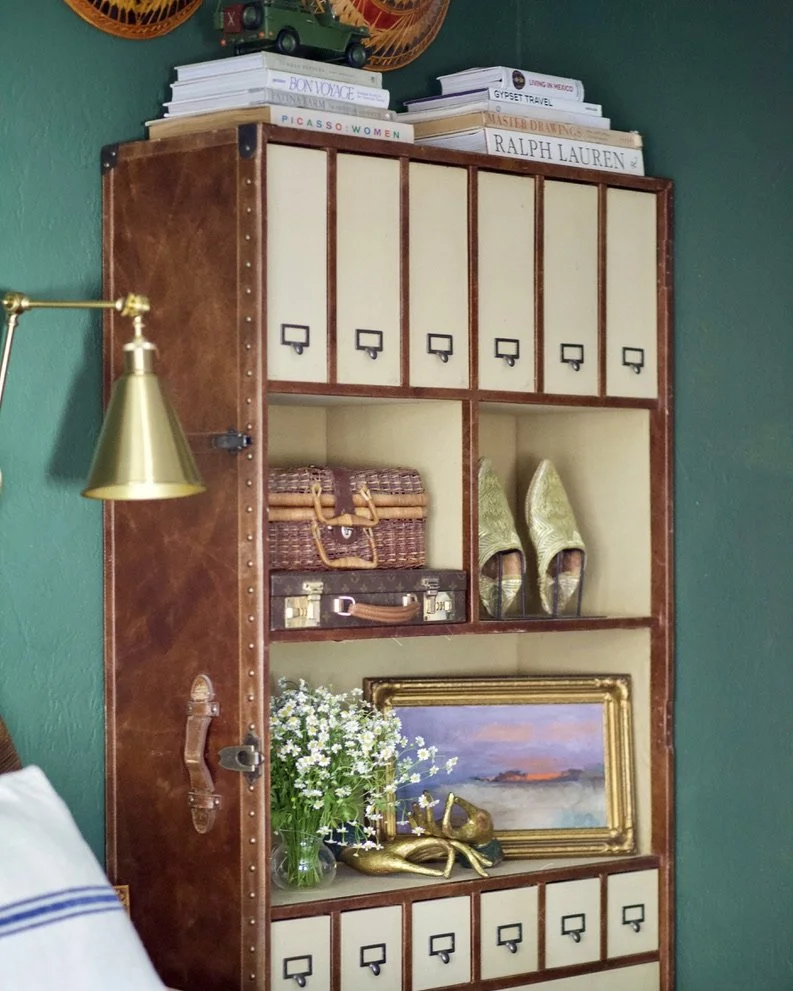 Decorative bookcase with vintage and eclectic decor including books, a gold desk lamp, woven baskets, vintage suitcase, gold shoes, a framed art piece, a bouquet of white flowers, and a golden swan sculpture, set against a green wall.