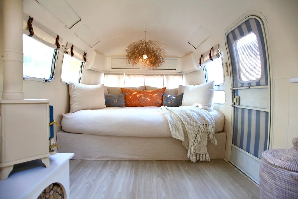 Interior of a cozy caravan or tiny home with a built-in bed surrounded by windows, decorated with pillows and a blanket, with a chandelier hanging from the ceiling.