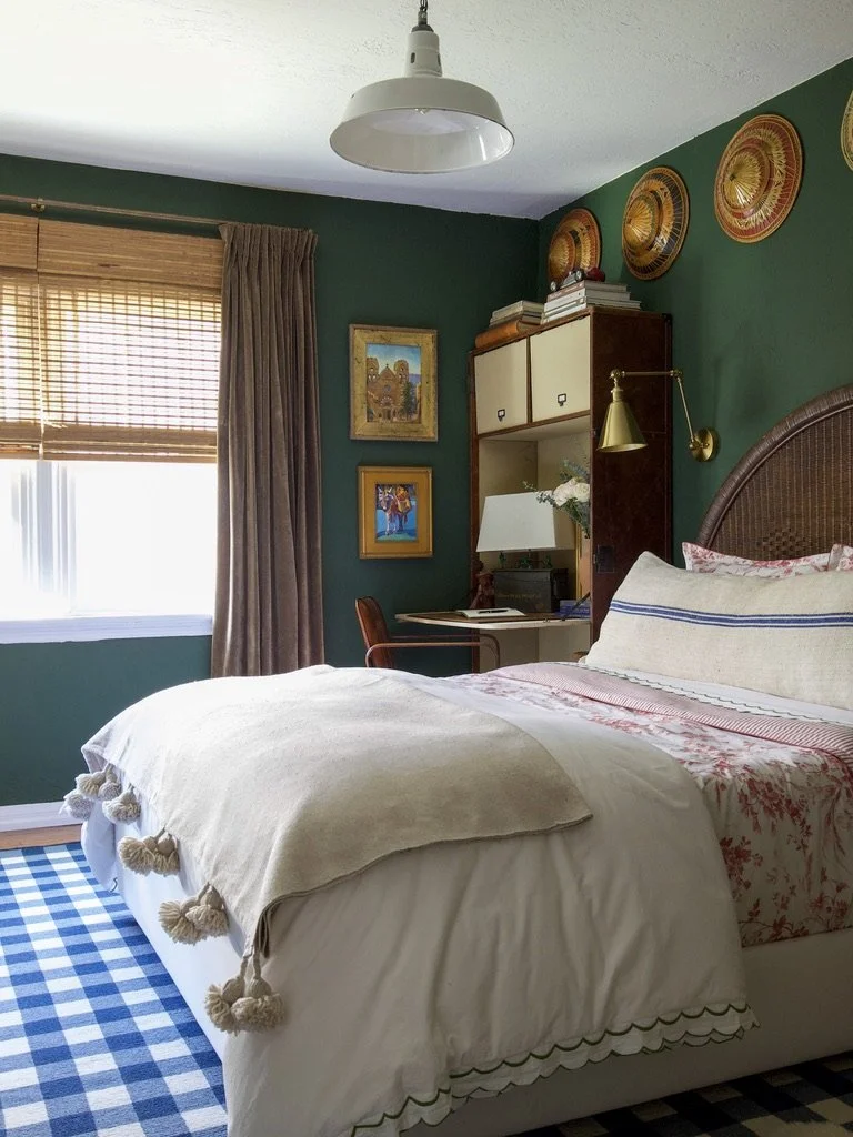 Cozy bedroom with green walls, a bed with patterned pillows, a wicker headboard, and a bedside wall-mounted lamp. A wood bookshelf, framed artwork, and a desk with a chair are visible near the window with bamboo blinds and beige curtains.
