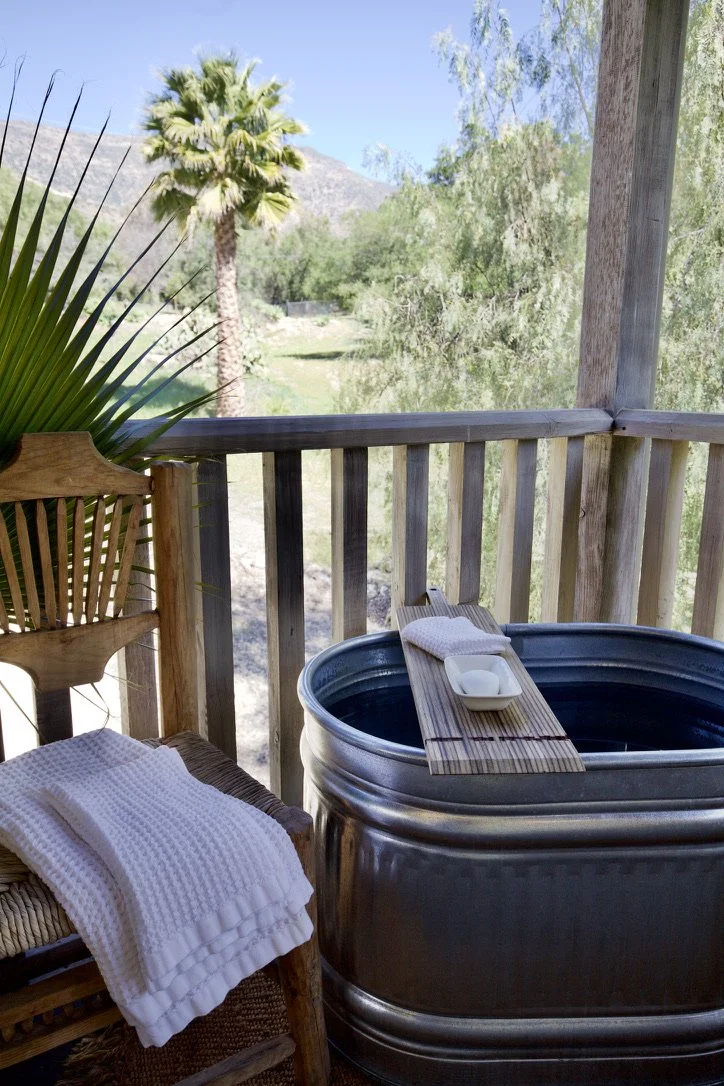 A wooden porch with a metal bathtub, a small wooden tray with soap and a towel, a wicker chair with a white towel, and a view of palm trees and mountains in the distance.