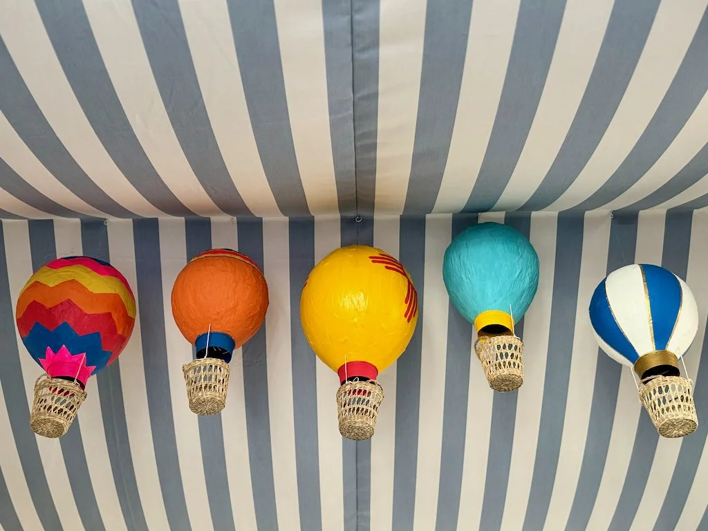 Five colorful paper lanterns resembling hot air balloons hanging from a ceiling with blue and white stripes.