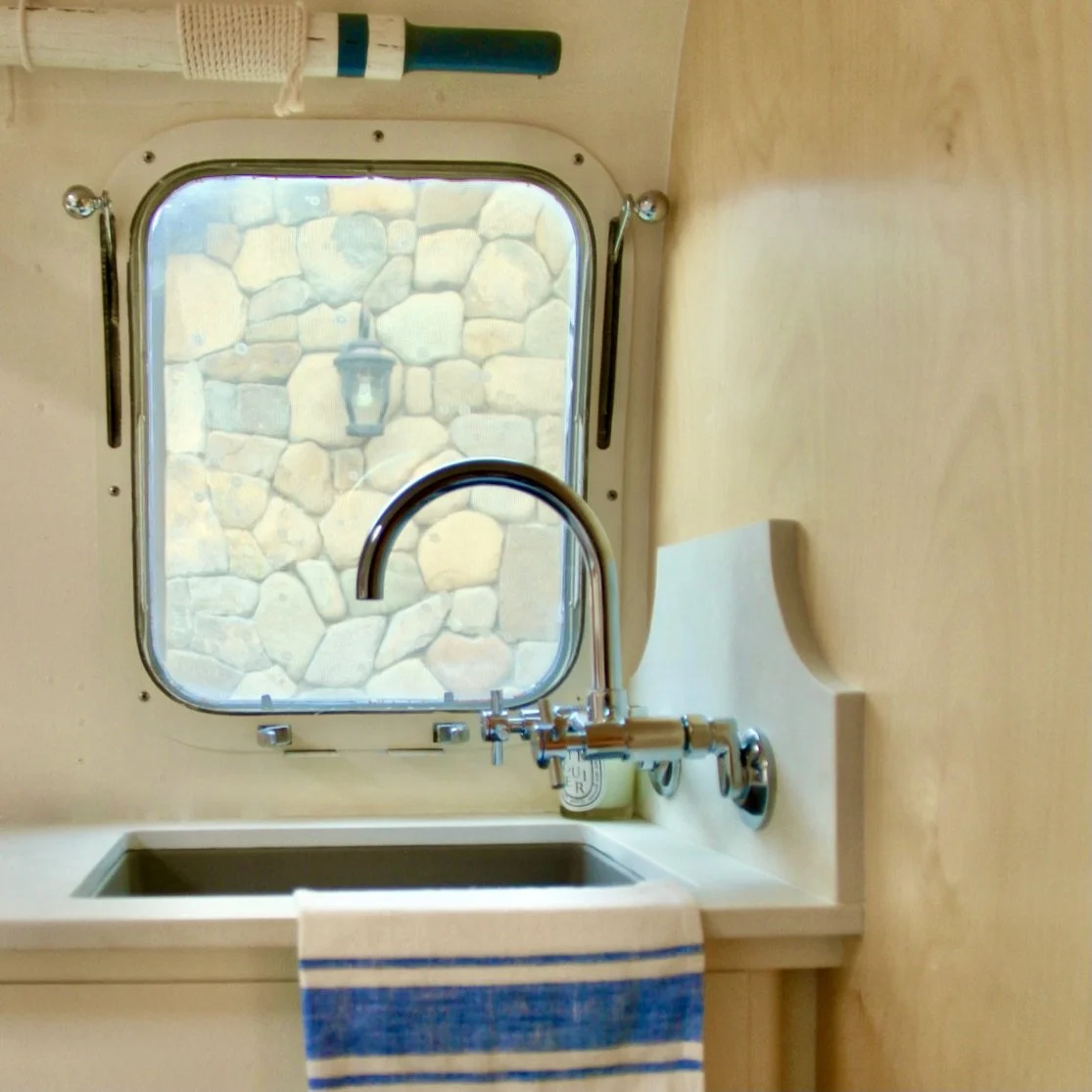 A small kitchen sink with a chrome faucet, a cup with utensils, a striped towel, a window with a stone wall outside, and a lamp hanging outside.