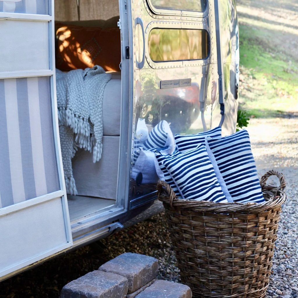 Inside a camper trailer parked outdoors, with a bed draped in a chunky white blanket visible through the open door. Outside, a basket filled with blue and white striped pillows sits on the gravel ground near the trailer.