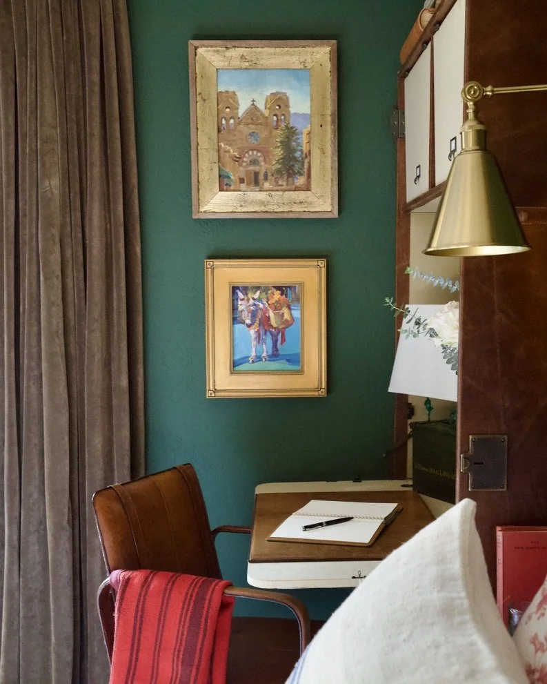 A cozy corner of a room with a small wooden desk, a brown leather chair with a red striped towel, and two framed paintings on a teal wall. A gold wall lamp is mounted on a wooden partition, and a white lamp with green foliage decoration is partially 