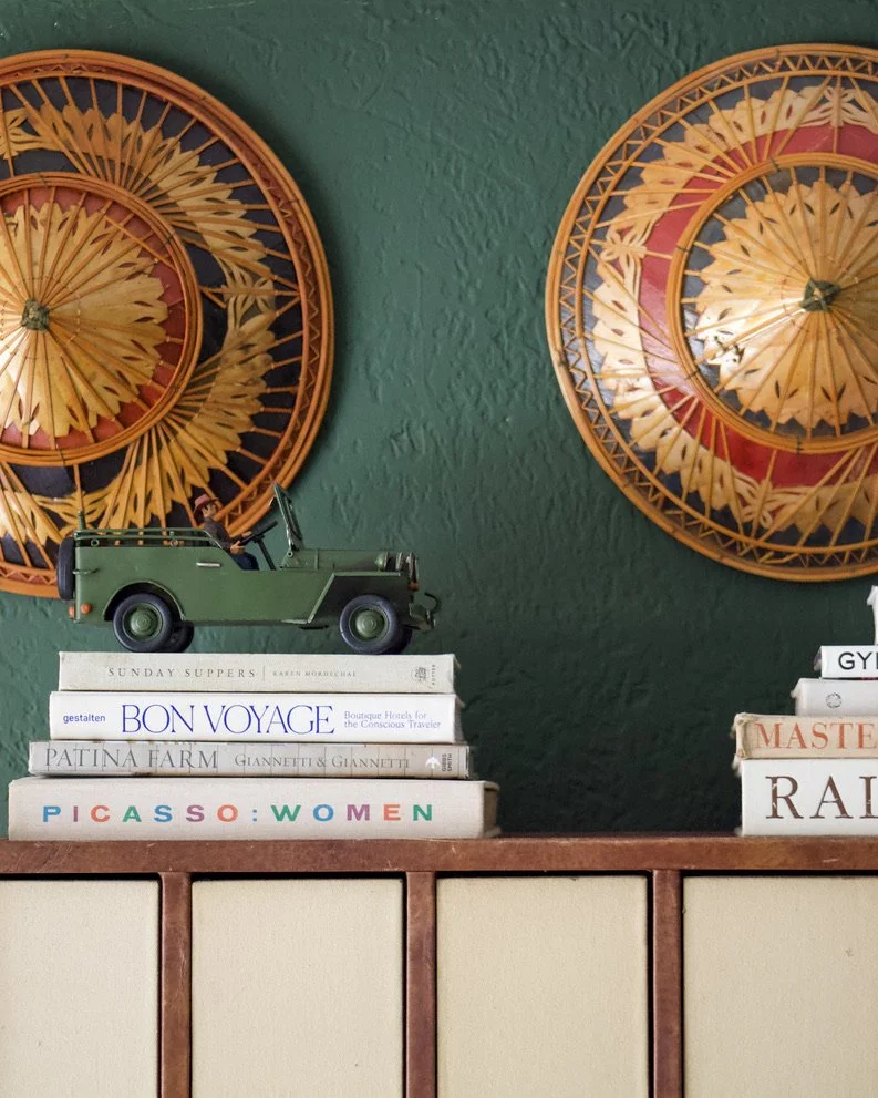 Decorative wall with two round, woven, yellow and orange wall hangings, with a green vintage toy jeep on top of a stack of books on a wooden cabinet.