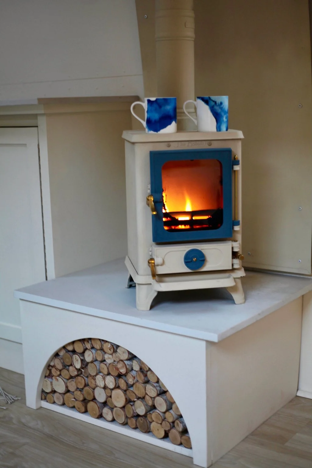 A small white wood stove with a blue door, burning fire inside, sitting on a white wooden stand with stacked firewood underneath. There are two white mugs with blue watercolor-like designs placed on top of the stove.