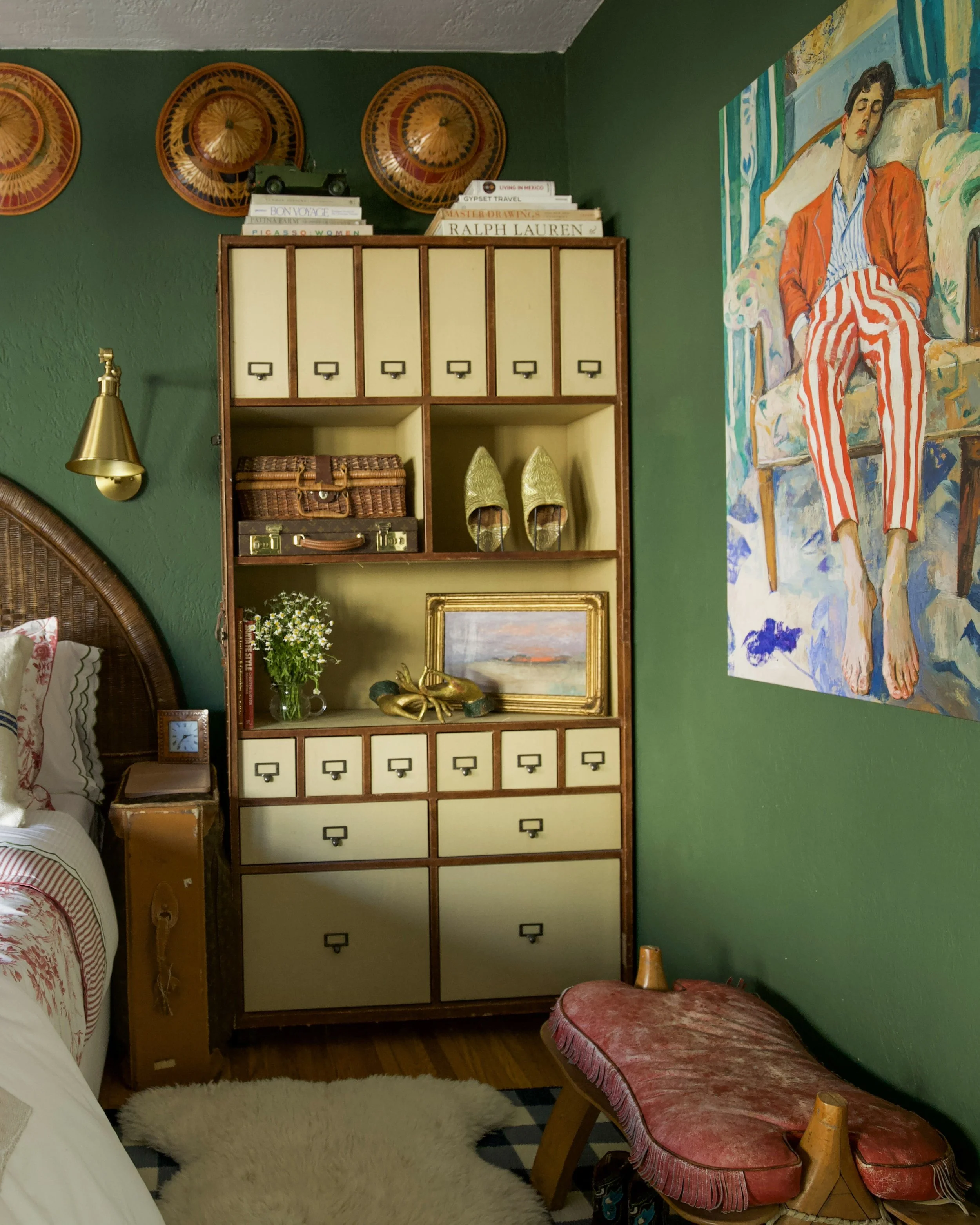 Interior of a bedroom with a vintage wooden cabinet, a framed painting, decorative shoes, and a large colorful painting of a woman on a green wall.