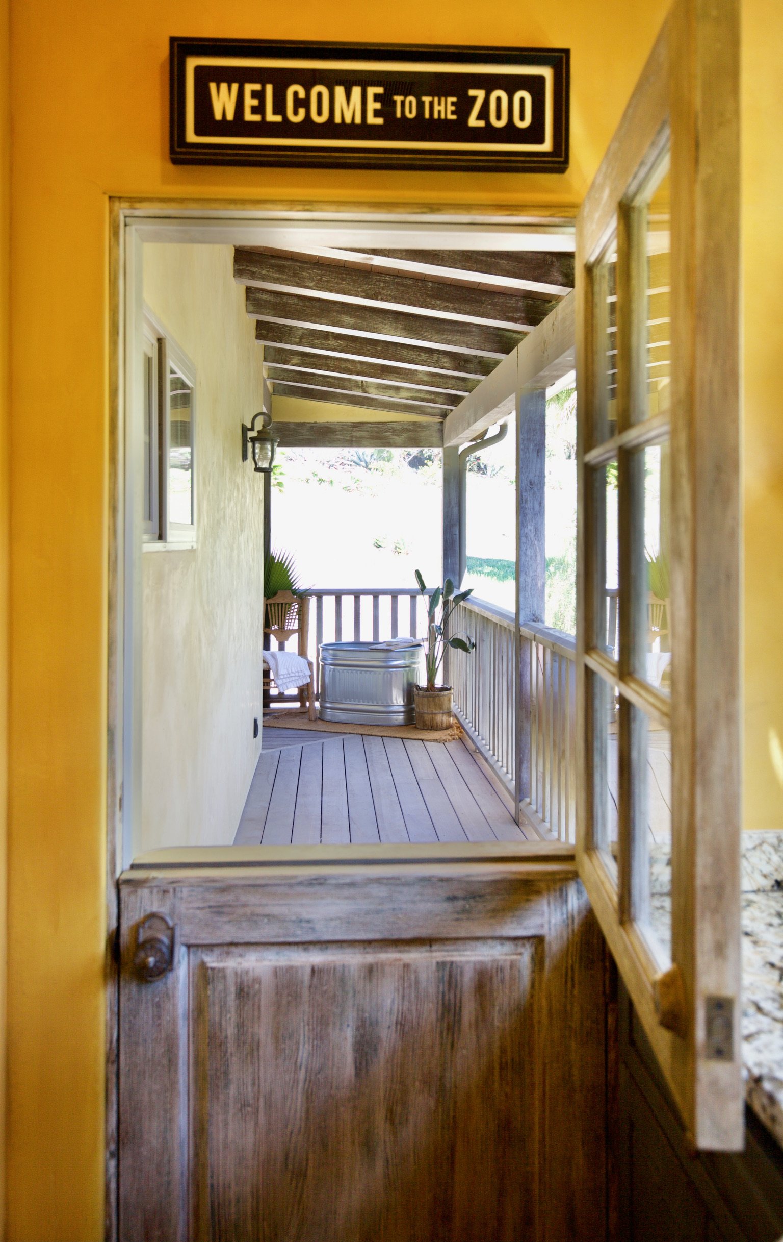 View through a wooden door into a yellow house with a sign that says "Welcome to the Zoo". Outside, on the porch, there are chairs, a potted plant, and a metal tub.