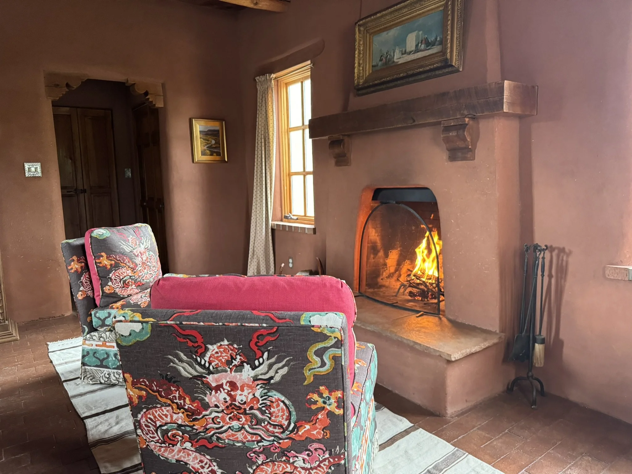 A cozy living room with a cozy fireplace with a fire burning. There are two chairs with colorful dragon-themed upholstery, facing the fireplace. A window with curtains allows natural light into the room. There are framed paintings on the walls, and a