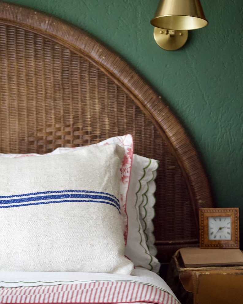 Close-up of a bed with a wicker headboard, decorative pillows, and a small bedside table with a clock, against a green textured wall with a brass wall sconce.