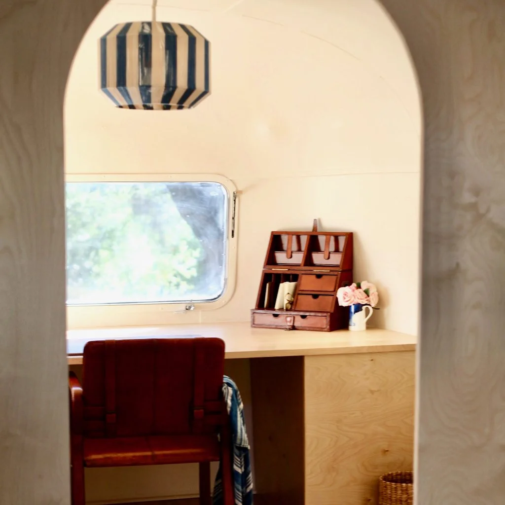 A cozy workspace inside a small wooden room with a desk, a wooden chair, a small wooden desk organizer, a vase with pink flowers, a window showing greenery outside, and a striped pendant light hanging from the ceiling.
