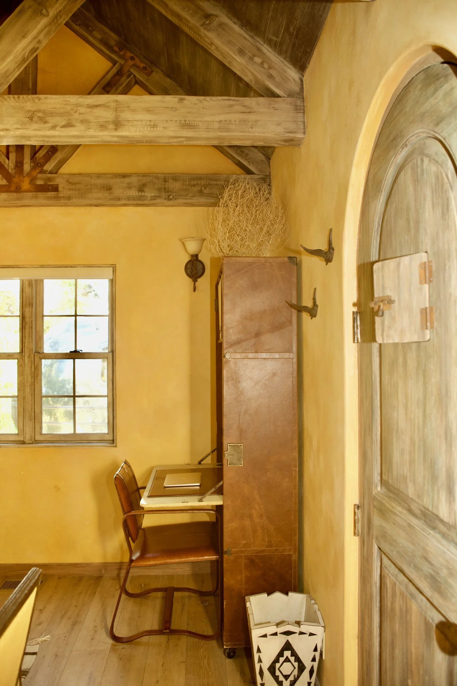 A cozy room with yellow walls, a window, a wooden desk with a chair, and a tall wooden cabinet. Hanging above the cabinet is a decorative dried branch sphere, and on the wall are two bird-shaped hooks. An arched wooden door is partially visible on th