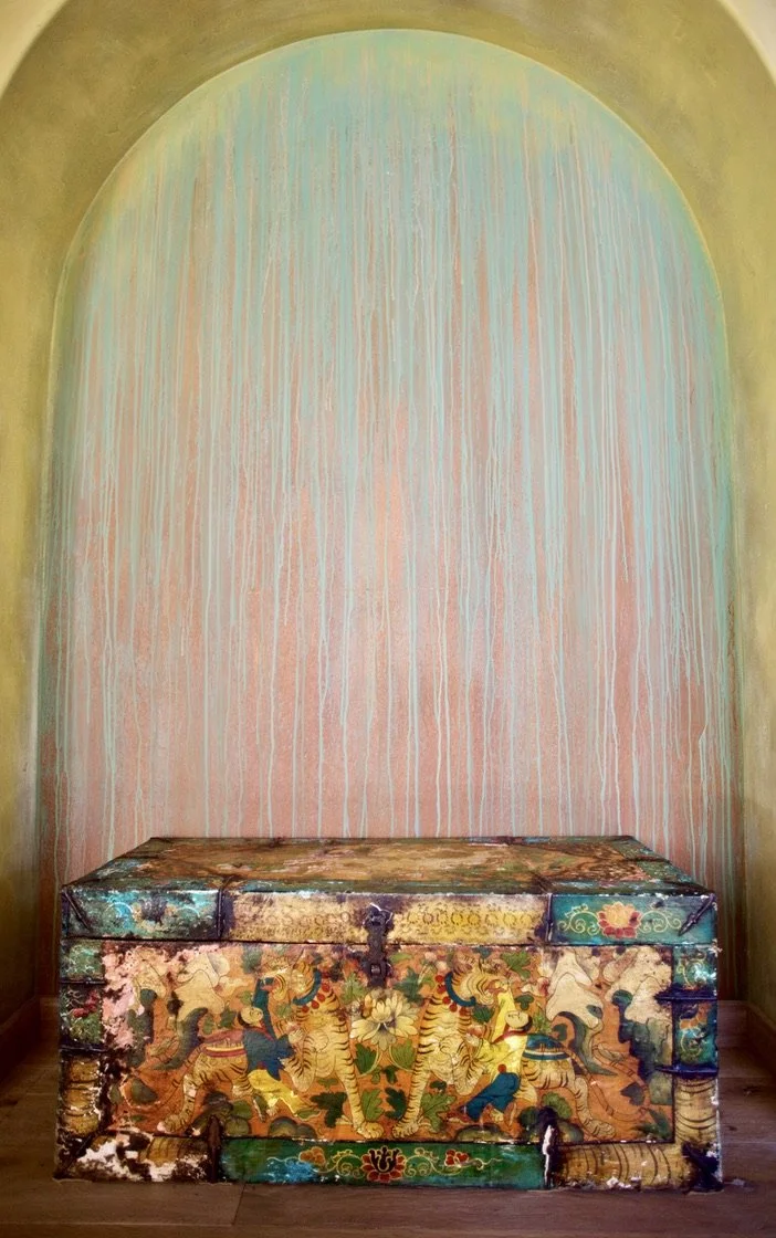 A decorative wooden chest with traditional Asian artwork, positioned against a pastel-colored arched wall with dripping paint design.