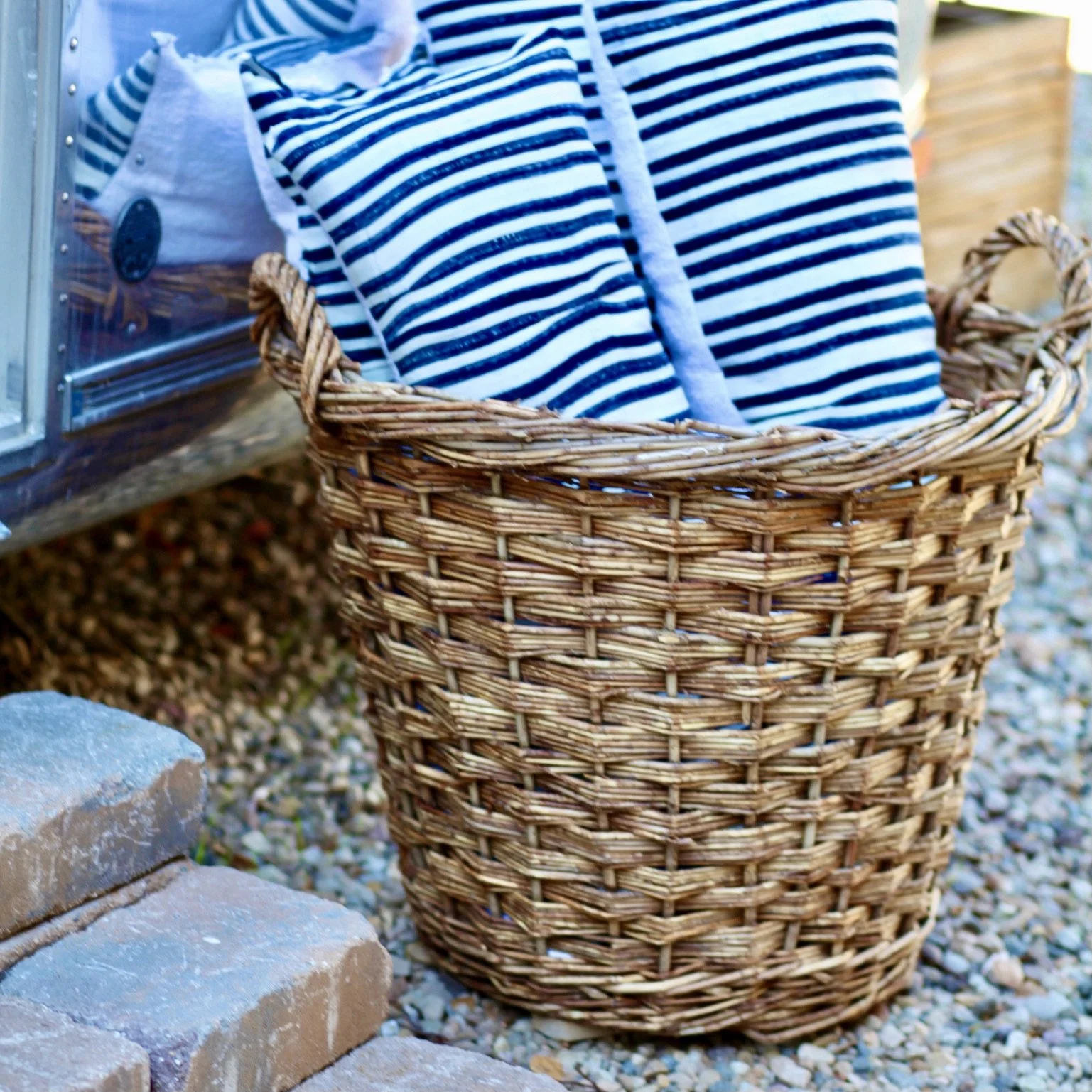 A woven wicker basket filled with folded navy and white striped clothing, placed on gravel next to a brick step.