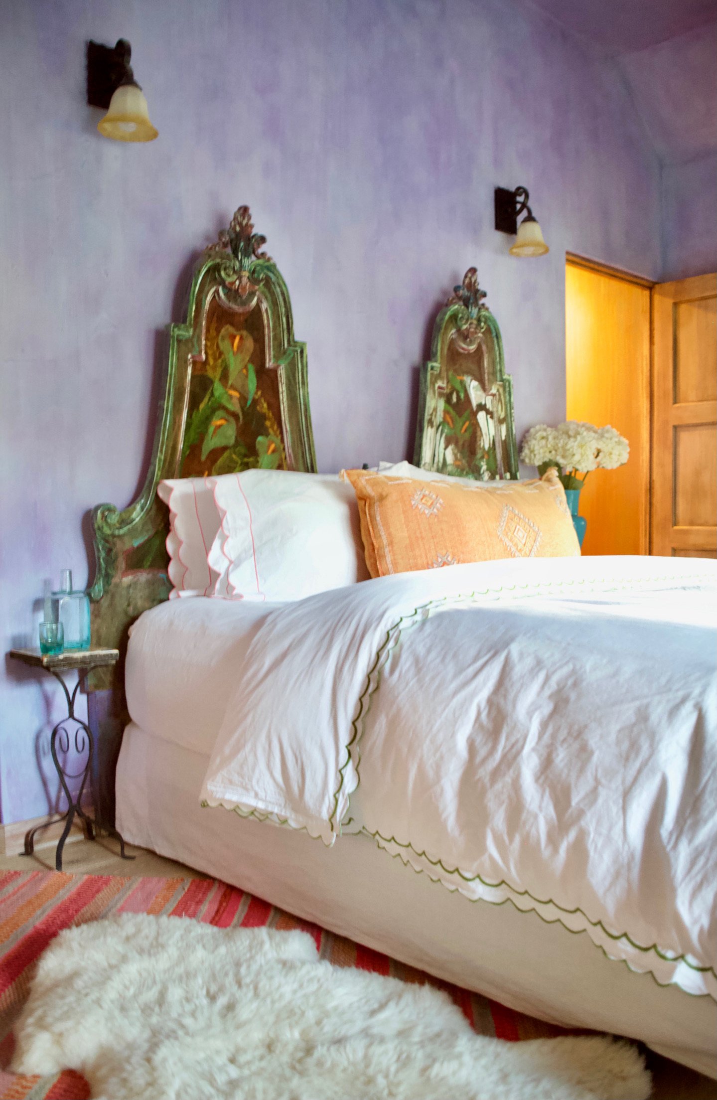 A cozy bedroom with a large bed featuring white bedding, decorative pillows, and an ornate headboard set against a light purple wall. A small side table with bottles and a turquoise vase with white flowers is next to the bed. A fluffy white rug is on