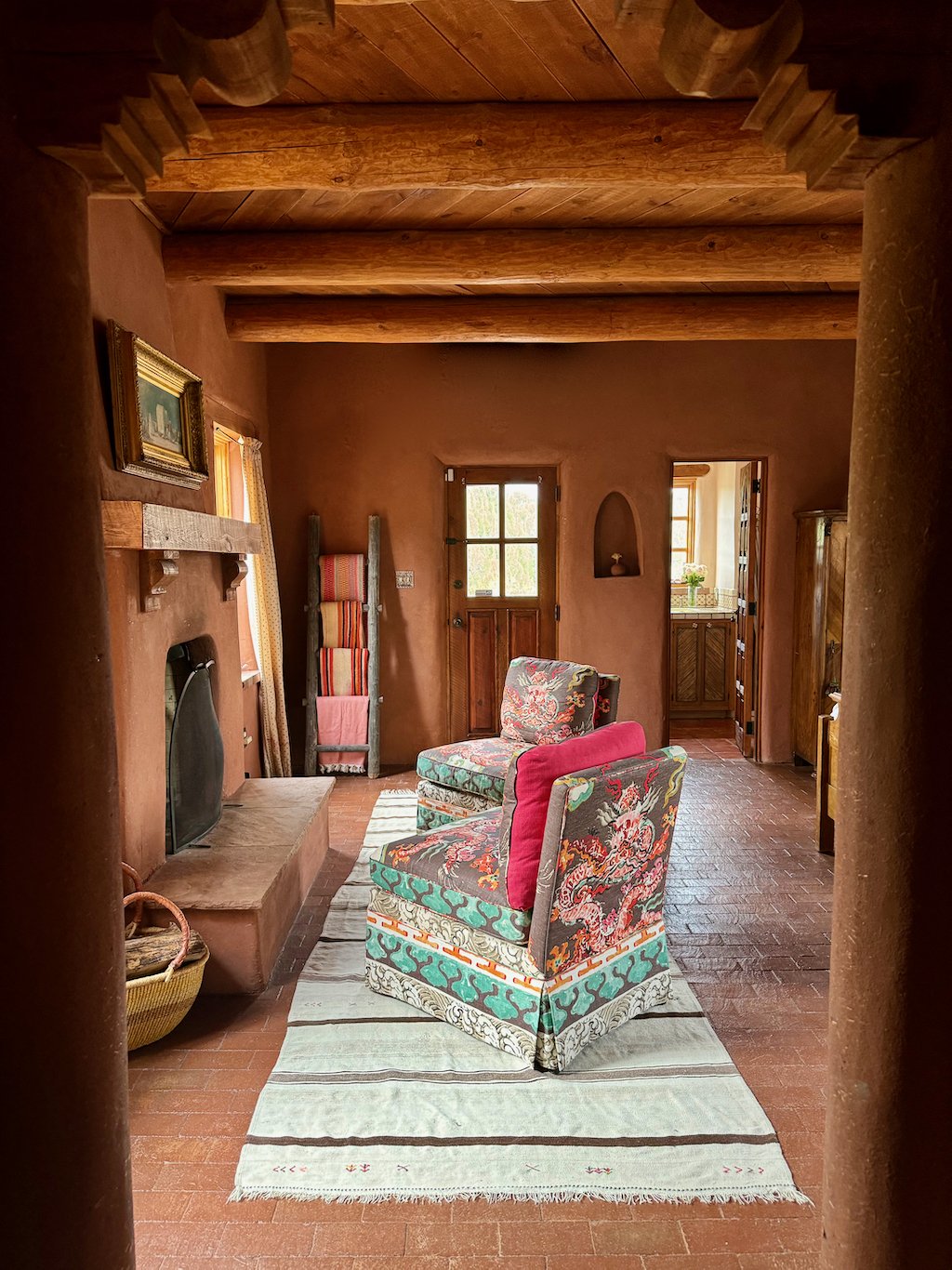 A cozy living room with wooden ceiling beams, a small fireplace, colorful patterned armchairs, a Rolled-up blanket, and a striped rug on a brick floor. There is a wooden door and a small window letting in natural light.