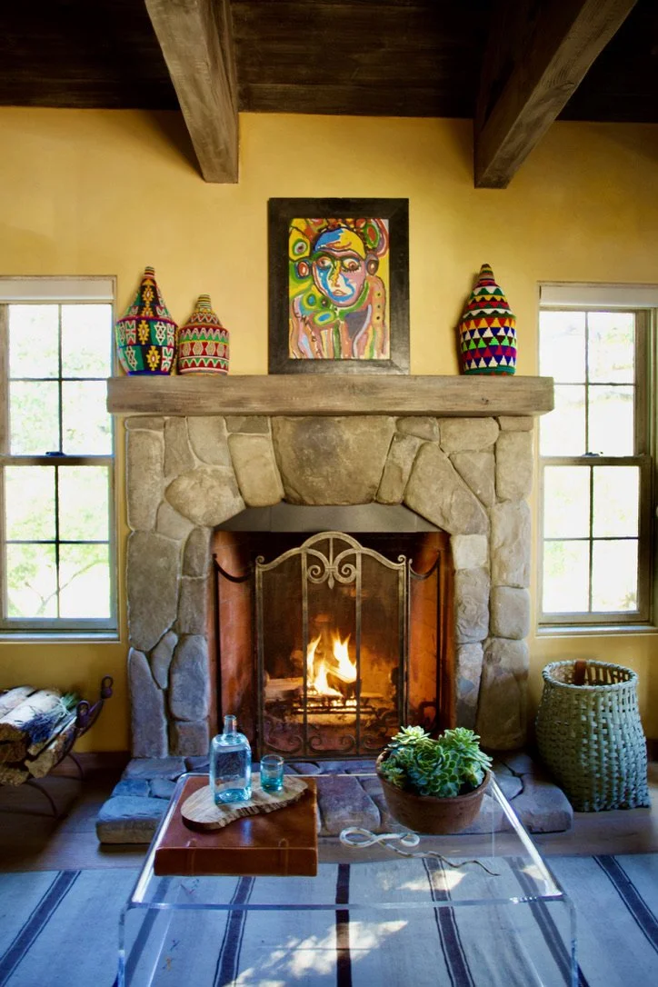 Living room with stone fireplace, wood mantel, colorful pottery on the mantle, wall art above fireplace, two windows with natural light, glass coffee table with water bottles, succulent plant, and a basket on the right side.
