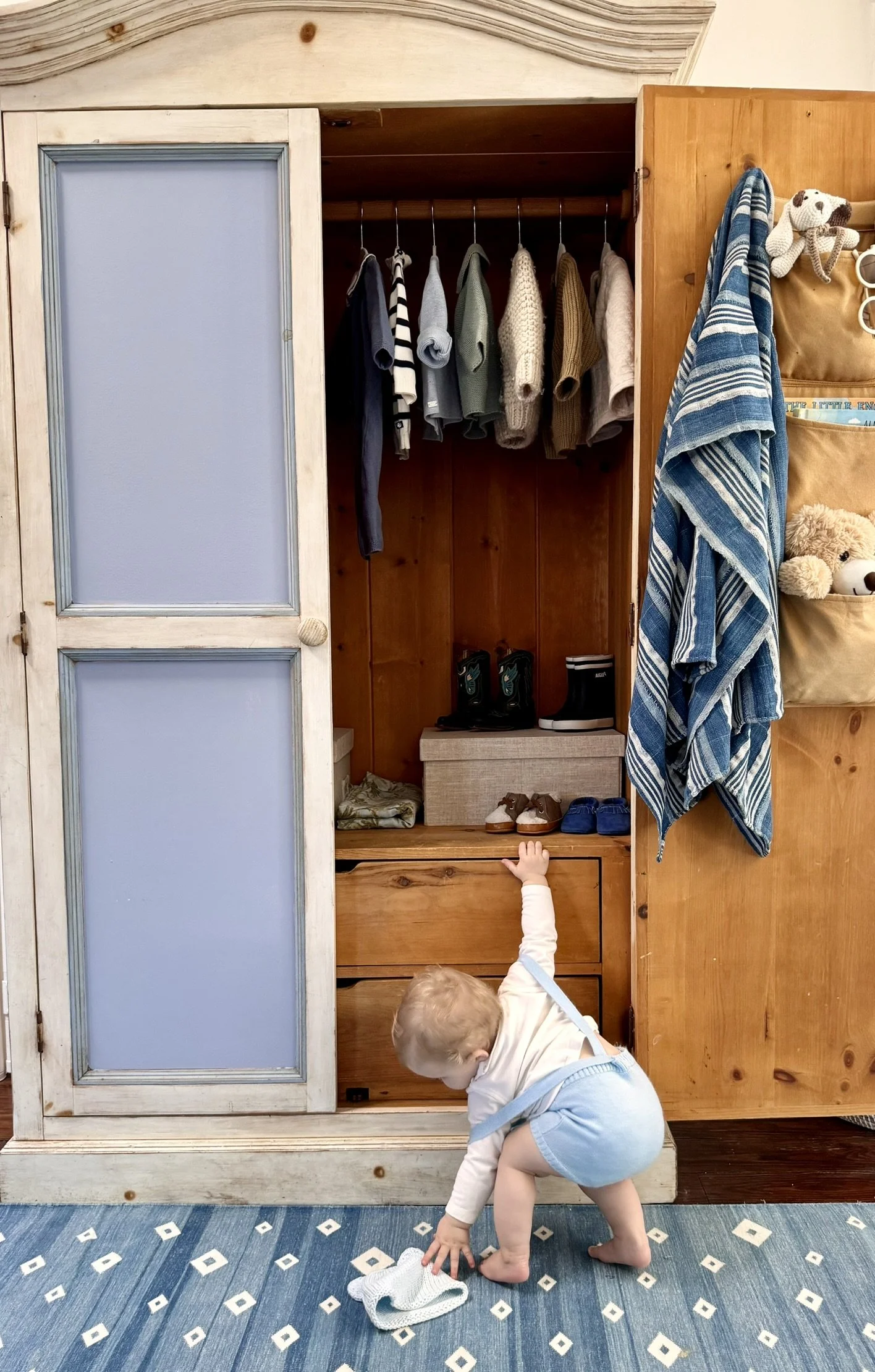 A toddler reaching into an open wooden closet with clothes hanging inside, shoes on a shelf, and a stuffed animal hanging on a nearby pocket organizer, on a blue patterned rug.