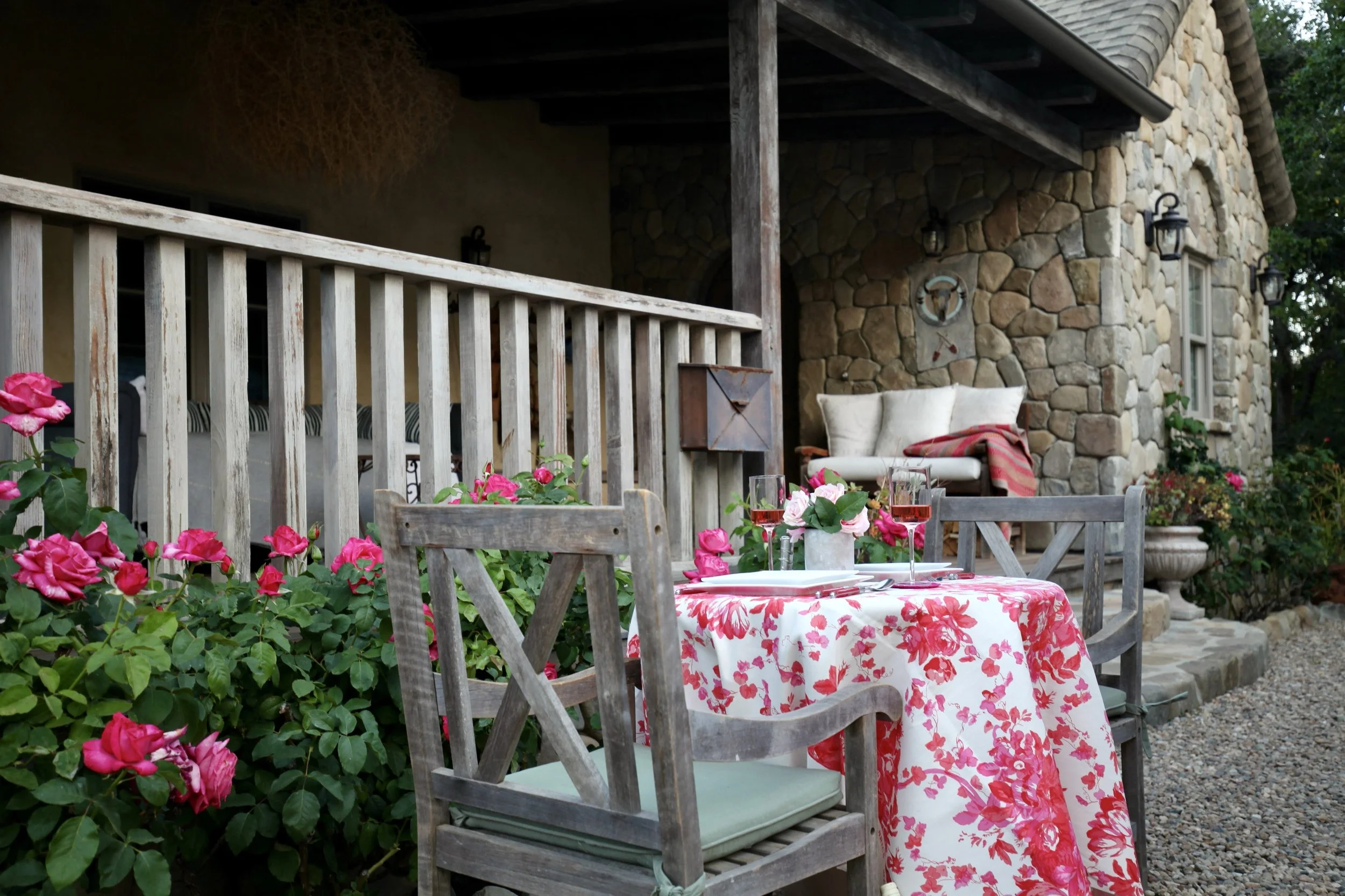 Outdoor patio with wooden table and chairs, pink flowers, and a stone house with porch swing and wall-mounted lamps.