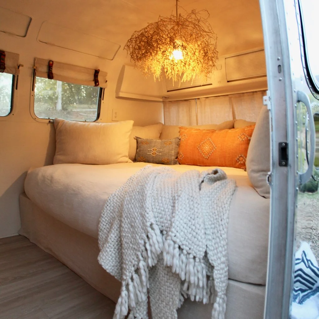 Cozy interior of a camper with a cream-colored bed or couch, adorned with various decorative pillows and a chunky knit blanket. A window is visible with a view of trees outside, and a warm hanging light fixture made of twigs or branches provides ambi