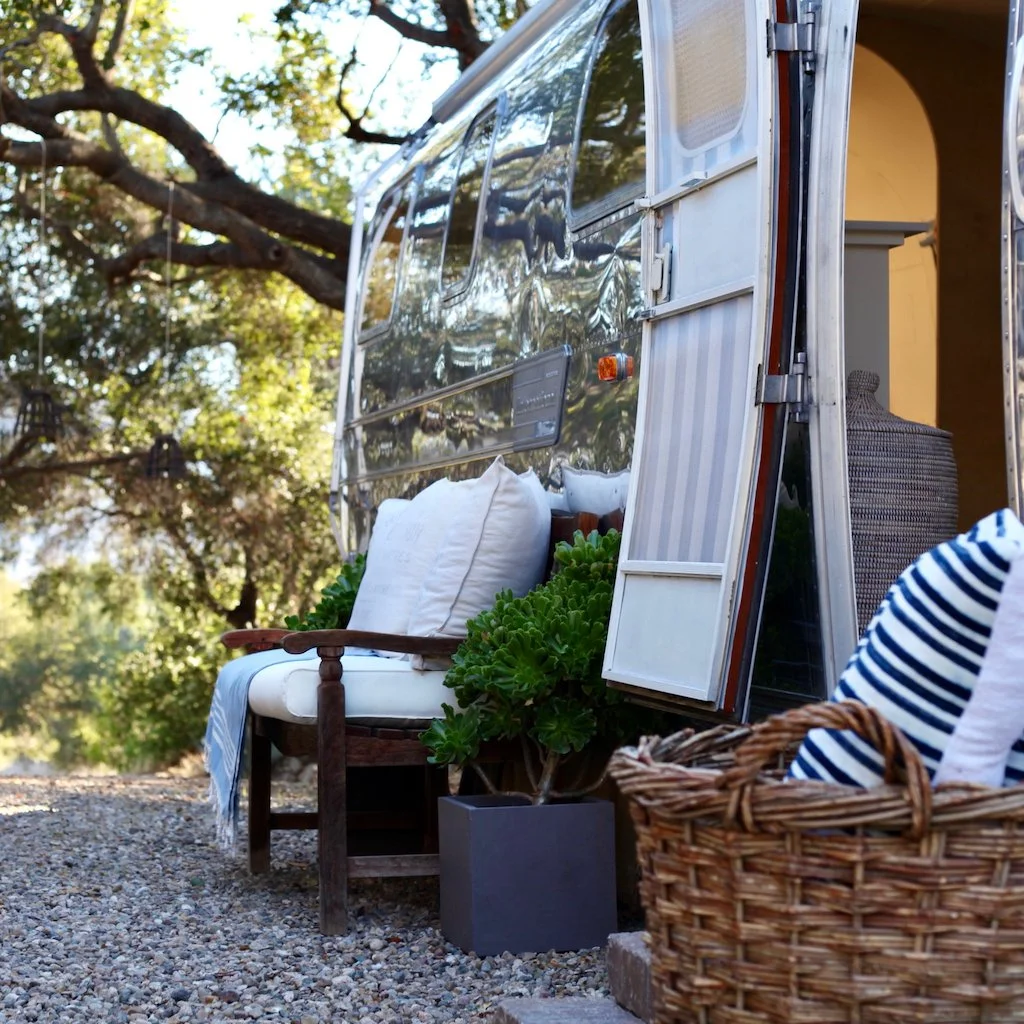 Outdoor scene featuring a shiny vintage Airstream travel trailer with a door open, revealing a cozy interior. In front of the trailer, there's a wooden bench with white cushions, a potted succulent plant, and a woven basket with striped fabric. The s