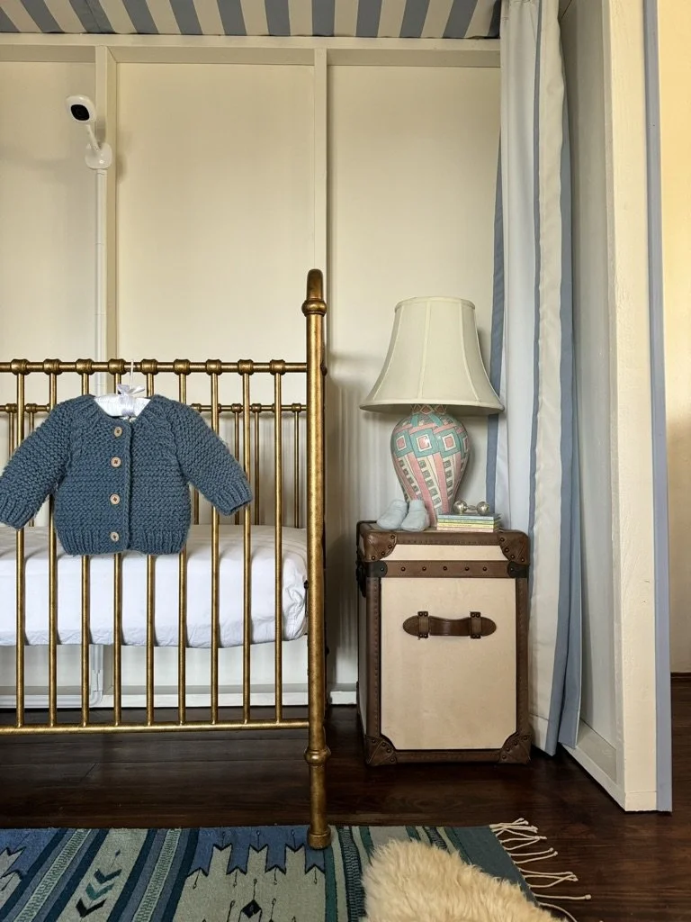 Child's room with a brass bed, a blue knitted sweater hanging on the bed, a vintage-style lamp on a small trunk, and a security camera in the upper left corner.