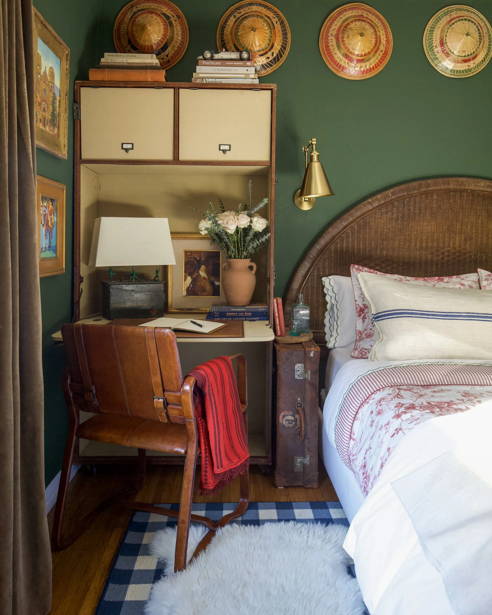 A cozy bedroom corner with a wooden desk, a brown leather chair, a bed with a woven headboard, colorful wall decorations, a desk lamp, flowers in a vase, books, and personal items.