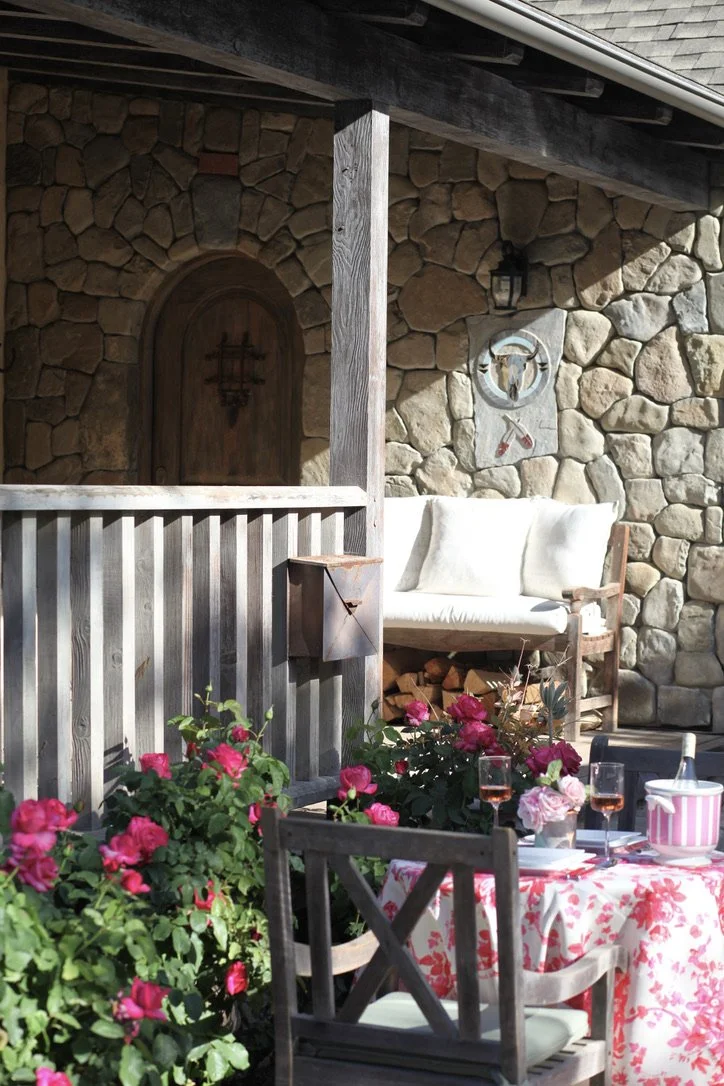 An outdoor seating area with a table covered in a pink floral tablecloth, pink roses, and two glasses of rosé wine. There are wooden chairs around the table, and a white cushioned bench against a stone wall. The stone wall has a decorative shield and