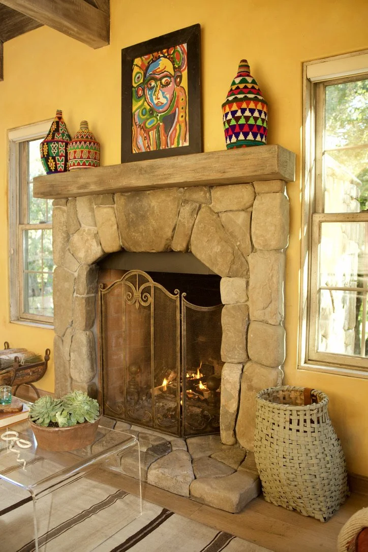 Living room with yellow walls, stone fireplace, colorful artwork above mantle, and decorative vases. Two windows with natural light, a woven basket beside the fireplace, and a clear table with a plant in the foreground.
