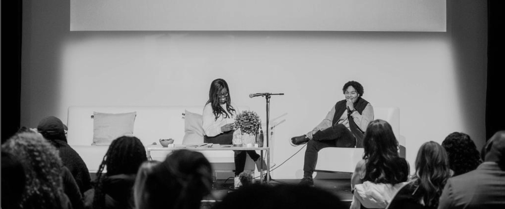 Two women sit on stage during a panel discussion or interview, facing an audience. One woman is holding a mug and smiling, while the other is speaking into a microphone and leaning back. The stage is decorated with pillows and a flower arrangement, with a large blank screen behind them.