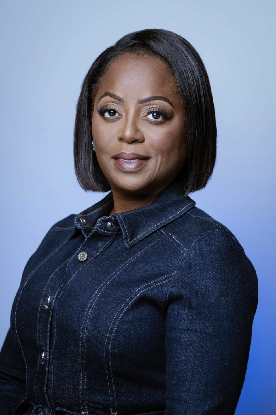 A woman with shoulder-length black hair, wearing a dark denim jacket, looking at the camera against a light blue background.