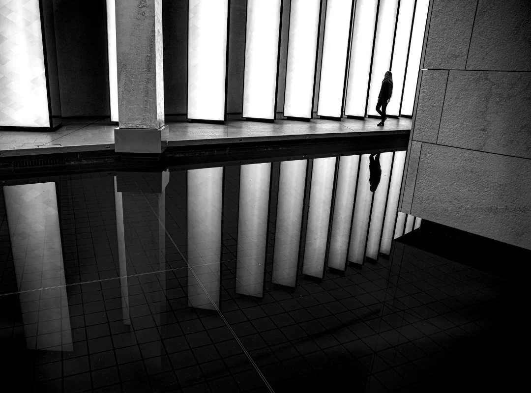 A person walking along a corridor with illuminated vertical panels on the wall, and their reflection visible in a reflective black floor.