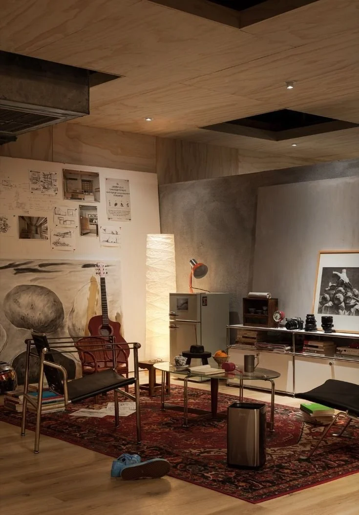 Cozy living room with a red Persian rug, a guitar leaning against the wall, a black and chrome chair, a red chair, a small side table with items, a tall white paper lamp, a white cabinet with a desk lamp, a framed photo, various camera lenses on a white console, and scattered papers and books.
