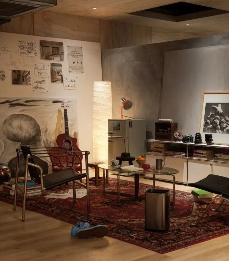 Cozy living room with modern and vintage decor, including a red guitar, glass coffee tables, a black lounge chair, a rug, and various art pieces and shelves on the wall.