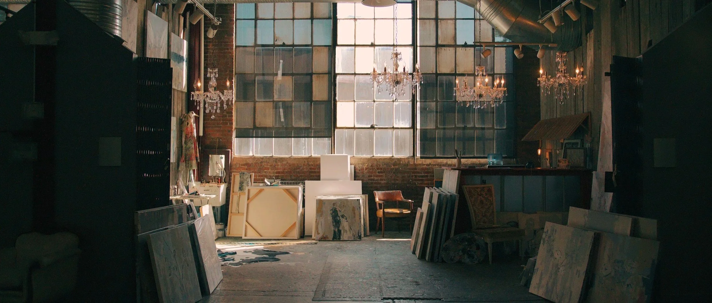 An art studio with large brick walls and tall windows. Several chandeliers hang from the ceiling, and various paintings and frames are stacked and leaning against the walls and furniture. The space has a vintage and creative atmosphere.
