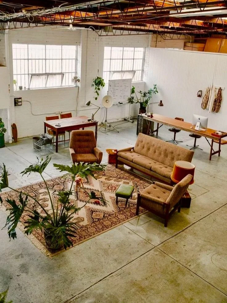 An industrial-style office space with vintage furniture, a rug, potted plants, a whiteboard, desks, and windows with natural light.