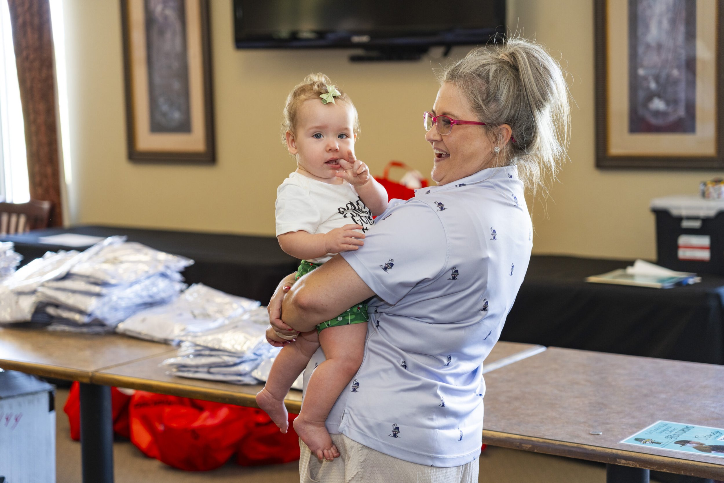 A woman with glasses and gray hair holding a young girl with a bow in her hair. The woman smiles while the girl touches her finger to her lips. There are tables with folded clothes in the background.